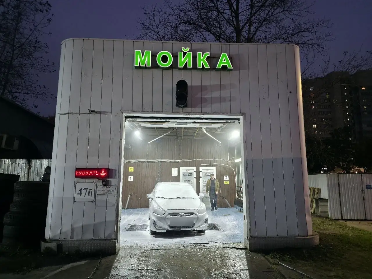 Въезд в мойку ночью: белое здание с зелёной вывеской «МОЙКА», в открытом боксе машина в пене